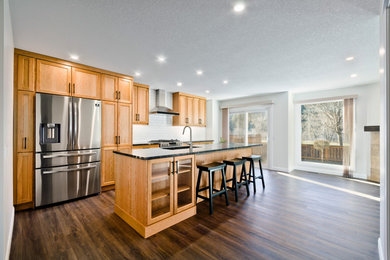 Mid-sized elegant single-wall vinyl floor and brown floor kitchen photo in Calgary with an undermount sink, shaker cabinets, medium tone wood cabinets, granite countertops, white backsplash, subway tile backsplash, stainless steel appliances, an island and black countertops