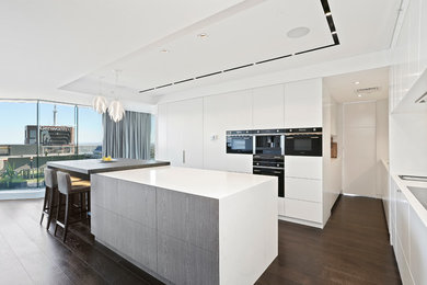 Photo of a large contemporary l-shaped kitchen pantry in Sydney with an undermount sink, flat-panel cabinets, white cabinets, solid surface benchtops, white splashback, stone slab splashback, black appliances, dark hardwood floors and with island.