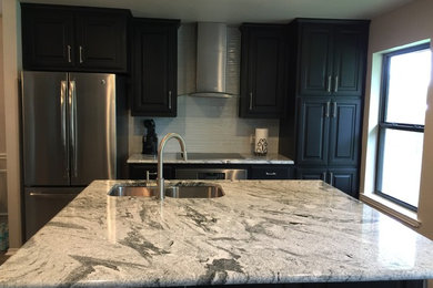 Large trendy l-shaped medium tone wood floor and brown floor eat-in kitchen photo in Dallas with a double-bowl sink, black cabinets, granite countertops, stainless steel appliances, an island, raised-panel cabinets, gray backsplash and matchstick tile backsplash