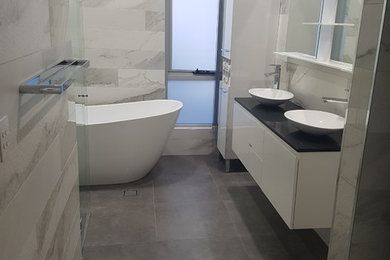 This is an example of a large modern master bathroom in Sydney with white cabinets, a freestanding tub, an open shower, a two-piece toilet, white tile, ceramic tile, white walls, concrete floors, engineered quartz benchtops, grey floor, a hinged shower door and black benchtops.
