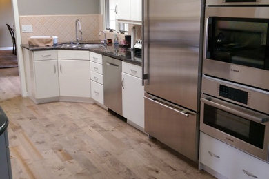 Photo of a mid-sized contemporary galley separate kitchen in San Francisco with a double-bowl sink, flat-panel cabinets, white cabinets, granite benchtops, beige splashback, ceramic splashback, stainless steel appliances, light hardwood floors and brown floor.