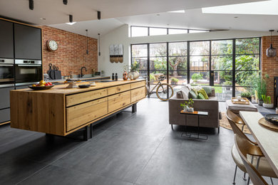Design ideas for a large contemporary grey and black single-wall open plan kitchen in Sussex with a built-in sink, flat-panel cabinets, distressed cabinets, quartz worktops, multi-coloured splashback, brick splashback, black appliances, ceramic flooring, an island, grey floors, grey worktops, a vaulted ceiling and a feature wall.