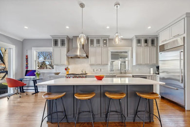 Example of a trendy medium tone wood floor kitchen design in Chicago with a farmhouse sink, recessed-panel cabinets, gray cabinets, solid surface countertops, white backsplash, stone tile backsplash, stainless steel appliances, an island and white countertops