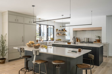 Photo of a large contemporary kitchen in Portland Maine with a farmhouse sink, recessed-panel cabinets, quartz benchtops, white splashback, ceramic splashback, panelled appliances, light hardwood floors, multiple islands and white benchtop.