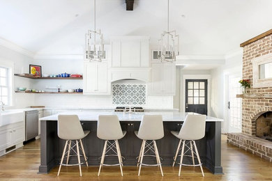 Example of a transitional l-shaped medium tone wood floor and brown floor kitchen design in Charleston with a farmhouse sink, shaker cabinets, white cabinets, white backsplash, stainless steel appliances, an island and white countertops