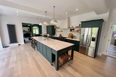 Photo of a large classic single-wall kitchen/diner in Dorset with a belfast sink, shaker cabinets, green cabinets, quartz worktops, white splashback, marble splashback, stainless steel appliances, light hardwood flooring, an island, brown floors and white worktops.