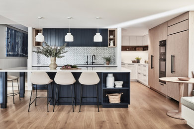 Photo of an expansive beach style l-shaped open plan kitchen in Sydney with a drop-in sink, shaker cabinets, blue cabinets, quartz benchtops, green splashback, porcelain splashback, black appliances, light hardwood floors, with island, beige floor and white benchtop.