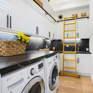 Foto de cuarto de lavado en L, actual, de tamaño medio, con armarios con paneles lisos, puertas de armario blancas, suelo de madera clara, lavadora y secadora juntas y encimeras negras