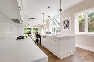 Inspiration for an expansive contemporary l-shaped open plan kitchen in Sydney with an undermount sink, shaker cabinets, white cabinets, solid surface benchtops, white splashback, subway tile splashback, stainless steel appliances, medium hardwood floors, with island, brown floor and white benchtop.