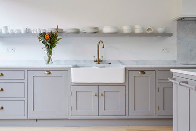 Inspiration for a medium sized contemporary l-shaped kitchen/diner in London with shaker cabinets, grey cabinets, composite countertops, white splashback, black appliances, an island, a belfast sink and medium hardwood flooring.