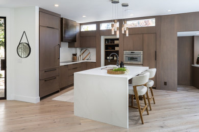 Example of a mid-sized trendy l-shaped light wood floor and brown floor eat-in kitchen design in San Francisco with a farmhouse sink, shaker cabinets, medium tone wood cabinets, quartz countertops, white backsplash, quartz backsplash, stainless steel appliances, an island and white countertops
