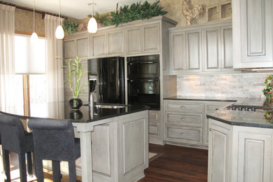 Kitchen - mid-sized traditional l-shaped medium tone wood floor kitchen idea in Minneapolis with a double-bowl sink, raised-panel cabinets, gray cabinets, granite countertops, gray backsplash, stone tile backsplash, black appliances and an island