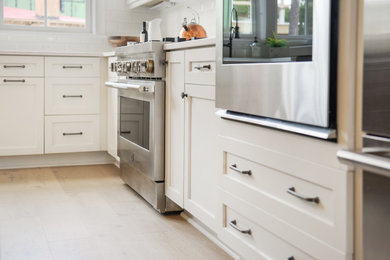 Open concept kitchen - large country l-shaped light wood floor and brown floor open concept kitchen idea in Atlanta with a farmhouse sink, shaker cabinets, white cabinets, quartz countertops, white backsplash, subway tile backsplash, stainless steel appliances, an island and white countertops