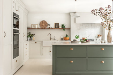 This is an example of a large classic l-shaped kitchen/diner in Surrey with a belfast sink, shaker cabinets, green cabinets, quartz worktops, white splashback, stone tiled splashback, black appliances, ceramic flooring, an island, beige floors and white worktops.