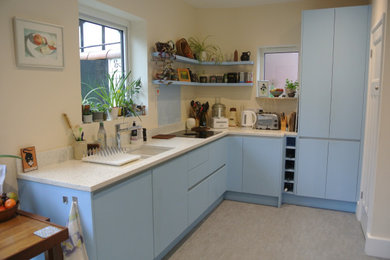 Photo of a mid-sized modern l-shaped separate kitchen in Hampshire with a drop-in sink, flat-panel cabinets, white cabinets, recycled glass benchtops, blue splashback, glass sheet splashback, black appliances and white benchtop.