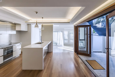 Photo of a mid-sized modern single-wall open plan kitchen in Sydney with quartz benchtops, stainless steel appliances, with island, an undermount sink, flat-panel cabinets, white cabinets and medium hardwood floors.