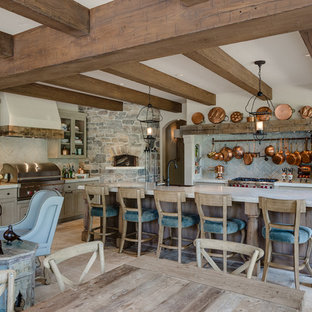 Diseño de cocina clásica, abierta, con armarios tipo vitrina, puertas de armario de madera clara, salpicadero beige, electrodomésticos de acero inoxidable y una isla