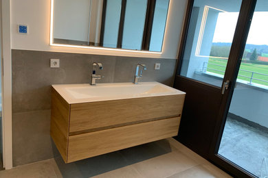 Mittelgroßes Modernes Badezimmer mit Kassettenfronten, hellen Holzschränken, grauen Fliesen, grauer Wandfarbe, Wandwaschbecken, Mineralwerkstoff-Waschtisch, weißer Waschtischplatte, Doppelwaschbecken und schwebendem Waschtisch