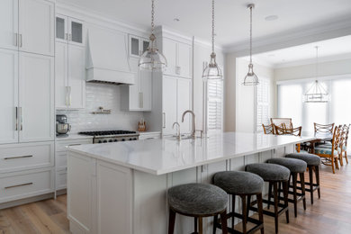Example of a mid-sized beach style medium tone wood floor and beige floor kitchen design in Miami with a farmhouse sink, shaker cabinets, white cabinets, quartz countertops, white backsplash, stainless steel appliances, an island, white countertops and ceramic backsplash