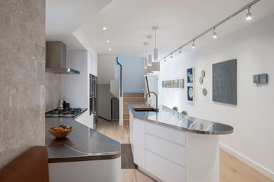 Modelo de cocina minimalista con armarios con paneles lisos, puertas de armario blancas, encimera de acero inoxidable, suelo de madera clara y una isla