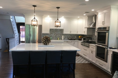 Example of a large transitional l-shaped dark wood floor and brown floor enclosed kitchen design in New York with recessed-panel cabinets, an island, a farmhouse sink, white cabinets, quartz countertops, gray backsplash, glass sheet backsplash and stainless steel appliances