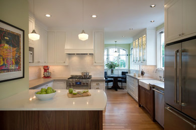 Mid-sized elegant l-shaped medium tone wood floor and brown floor enclosed kitchen photo in Portland with a farmhouse sink, shaker cabinets, white cabinets, quartz countertops, white backsplash, subway tile backsplash, stainless steel appliances and a peninsula