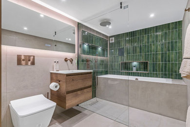 This is an example of a medium sized contemporary family bathroom in London with light wood cabinets, a built-in bath, a walk-in shower, a wall mounted toilet, green tiles, ceramic tiles, ceramic flooring, a built-in sink, beige floors, a feature wall, a single sink and a floating vanity unit.