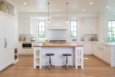 Example of a classic light wood floor kitchen design in Los Angeles with a farmhouse sink, shaker cabinets, white cabinets, white backsplash, subway tile backsplash, stainless steel appliances and an island
