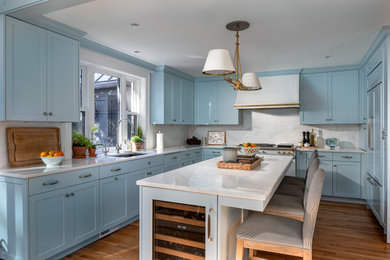 This is an example of a mid-sized l-shaped eat-in kitchen in DC Metro with a drop-in sink, recessed-panel cabinets, blue cabinets, marble benchtops, white splashback, marble splashback, white appliances, medium hardwood floors, with island, brown floor and white benchtop.