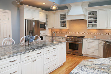 Photo of a large traditional u-shaped eat-in kitchen in New York with an undermount sink, raised-panel cabinets, white cabinets, granite benchtops, beige splashback, ceramic splashback, stainless steel appliances, medium hardwood floors, with island, brown floor and multi-coloured benchtop.