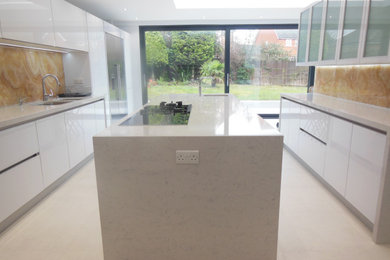 This is an example of a large modern galley kitchen/diner in London with a double-bowl sink, flat-panel cabinets, white cabinets, quartz worktops, yellow splashback, stone slab splashback, integrated appliances, porcelain flooring, an island, white floors and white worktops.