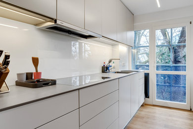 Zweizeilige, Kleine Moderne Küche mit Einbauwaschbecken, flächenbündigen Schrankfronten, Küchenrückwand in Weiß, Glasrückwand, schwarzen Elektrogeräten und braunem Holzboden in Nürnberg