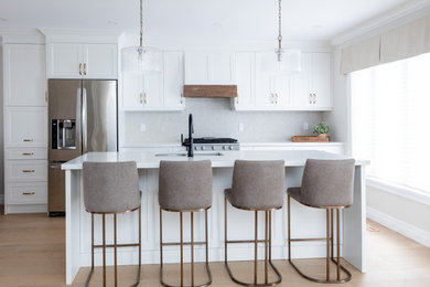 Mid-sized trendy single-wall light wood floor and brown floor kitchen photo in Toronto with an undermount sink, shaker cabinets, white cabinets, quartz countertops, white backsplash, ceramic backsplash, stainless steel appliances, an island and white countertops