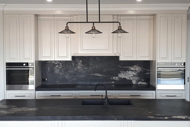 Inspiration for a large traditional galley open plan kitchen in Other with an undermount sink, recessed-panel cabinets, white cabinets, marble benchtops, black splashback, marble splashback, stainless steel appliances, vinyl floors, with island, brown floor and black benchtop.