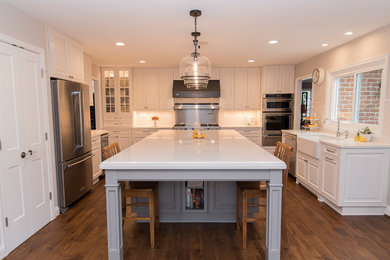 Inspiration for a large country u-shaped eat-in kitchen in Baltimore with a farmhouse sink, raised-panel cabinets, white cabinets, quartz benchtops, white splashback, ceramic splashback, stainless steel appliances, medium hardwood floors, with island, brown floor and white benchtop.