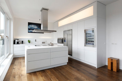 Große, Offene Moderne Küche in L-Form mit flächenbündigen Schrankfronten, weißen Schränken, Küchenrückwand in Weiß, Küchengeräten aus Edelstahl, dunklem Holzboden, Kücheninsel, Mineralwerkstoff-Arbeitsplatte, Einbauwaschbecken, Glasrückwand und braunem Boden in Hamburg
