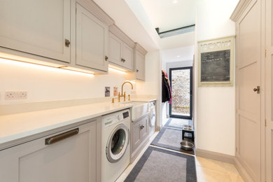 Photo of a medium sized contemporary galley utility room in Essex with a belfast sink, shaker cabinets, grey cabinets, engineered stone countertops, white walls, ceramic flooring, a side by side washer and dryer, beige floors and white worktops.