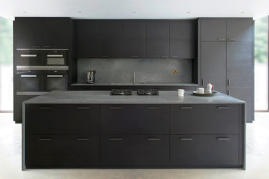 Photo of a large modern galley open plan kitchen in Sussex with an undermount sink, flat-panel cabinets, dark wood cabinets, solid surface benchtops, grey splashback, stone slab splashback, black appliances, concrete floors, with island, grey floor and grey benchtop.