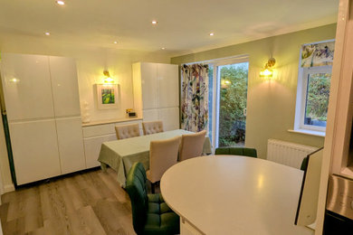 Photo of a medium sized contemporary grey and white l-shaped kitchen/diner in Surrey with a built-in sink, flat-panel cabinets, white cabinets, quartz worktops, green splashback, glass sheet splashback, black appliances, laminate floors, an island, grey floors, grey worktops and feature lighting.