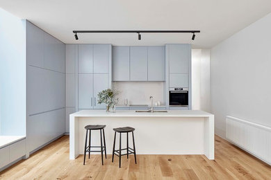 Inspiration for a mid-sized contemporary galley kitchen in Melbourne with a double-bowl sink, blue cabinets, quartz benchtops, white splashback, stone slab splashback, stainless steel appliances, light hardwood floors, with island, white benchtop, flat-panel cabinets and brown floor.