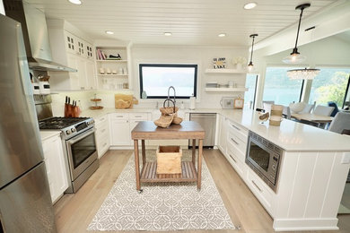 Design ideas for a mid-sized beach style u-shaped open plan kitchen with a farmhouse sink, recessed-panel cabinets, white cabinets, quartz benchtops, stainless steel appliances, light hardwood floors, a peninsula, brown floor and white benchtop.