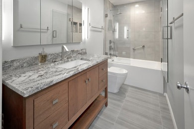 Small contemporary bathroom in DC Metro with flat-panel cabinets, medium wood cabinets, a shower/bathtub combo, a wall-mount toilet, gray tile, porcelain tile, blue walls, porcelain floors, an undermount sink, granite benchtops, grey floor, a hinged shower door and grey benchtops.