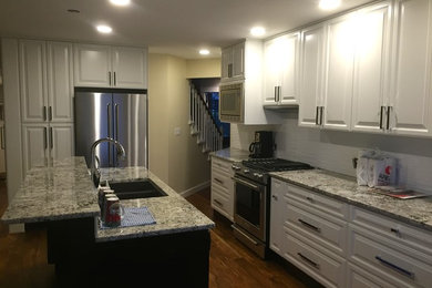 Open concept kitchen - mid-sized transitional l-shaped medium tone wood floor and brown floor open concept kitchen idea in Vancouver with raised-panel cabinets, white cabinets, granite countertops, white backsplash, ceramic backsplash, an island, a double-bowl sink and stainless steel appliances