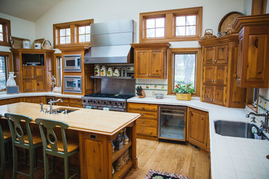 Large traditional u-shaped eat-in kitchen in Milwaukee with an undermount sink, raised-panel cabinets, medium wood cabinets, tile benchtops, multi-coloured splashback, stone tile splashback, light hardwood floors, with island, brown floor and stainless steel appliances.