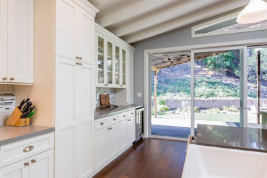 Mid-sized trendy l-shaped dark wood floor and brown floor open concept kitchen photo in Los Angeles with a farmhouse sink, raised-panel cabinets, white cabinets, wood countertops, gray backsplash, ceramic backsplash, stainless steel appliances, an island and gray countertops