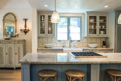 Example of a classic kitchen design in Santa Barbara with a farmhouse sink, glass-front cabinets, gray cabinets, beige backsplash and paneled appliances