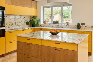 This is an example of a large retro l-shaped kitchen/diner in Kent with a submerged sink, flat-panel cabinets, yellow cabinets, terrazzo worktops, multi-coloured splashback, stone slab splashback, stainless steel appliances, medium hardwood flooring, an island, brown floors and multicoloured worktops.