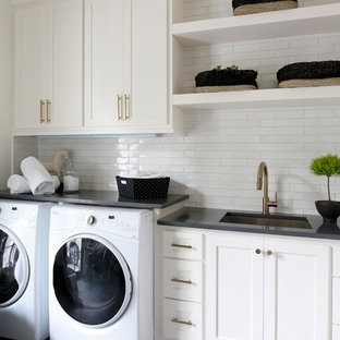 Modelo de cuarto de lavado lineal, marinero, con fregadero bajoencimera, armarios estilo shaker, puertas de armario blancas, paredes blancas, lavadora y secadora juntas, suelo gris y encimeras negras