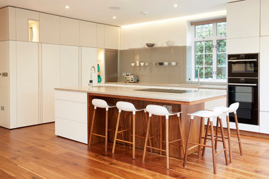 This is an example of a contemporary kitchen in London with flat-panel cabinets, white cabinets, beige splashback, glass sheet splashback, black appliances, medium hardwood floors and with island.