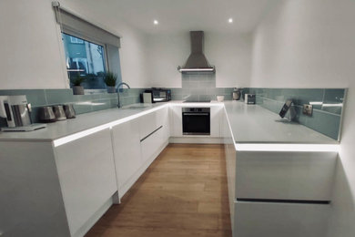 Small modern u-shaped kitchen/diner in Cornwall with an integrated sink, flat-panel cabinets, white cabinets, composite countertops, green splashback, cement tile splashback, stainless steel appliances, porcelain flooring, brown floors, white worktops and a drop ceiling.